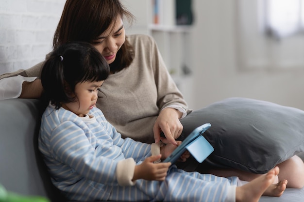 Foto menina asiática e mãe olham e tocam o tablet sentado no sofá menina jogando jogo usando a mesa em casa conceito de bebê da família de tecnologia
