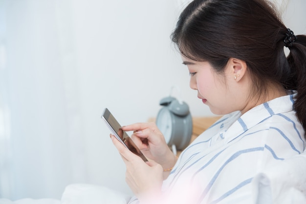 Menina asiática ela que relaxa e que joga o telefone celular esperto na cama, conceito em linha da compra.