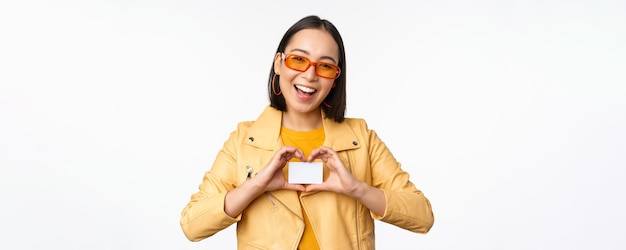 Menina asiática elegante e atraente em óculos de sol mostrando cartão de crédito e sorrindo em pé feliz contra o fundo branco do estúdio