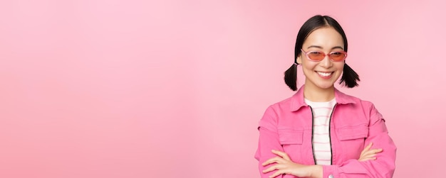 Menina asiática elegante em roupas cor de rosa e óculos de sol sorrindo e olhando feliz para a câmera em pé sobre o fundo do estúdio