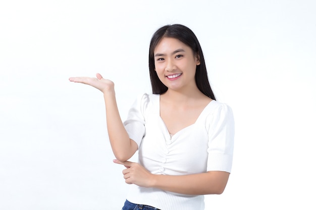 Menina asiática em cabelo comprido preto usa camisa branca e mostra apontar para apresentar algo sobre fundo branco.