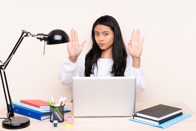 Menina asiática em um local de trabalho com um laptop na parede bege, fazendo o gesto de parada e desapontado