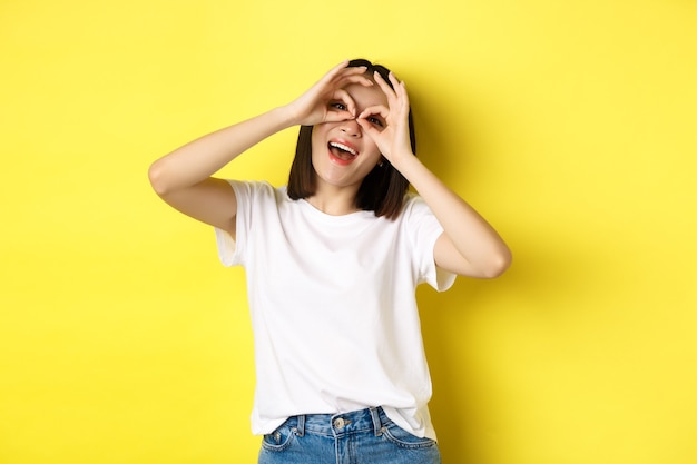 Menina asiática engraçada olhando através de binóculos de mão e sorrindo, stading sobre fundo amarelo.