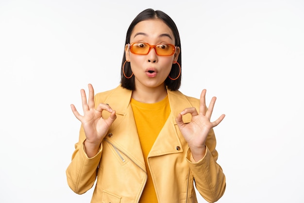 Menina asiática entusiasmada em óculos de sol mostra ok ok sinal de aprovação sorrindo e rindo aprova recomendar smth elogios e elogios em pé sobre fundo branco