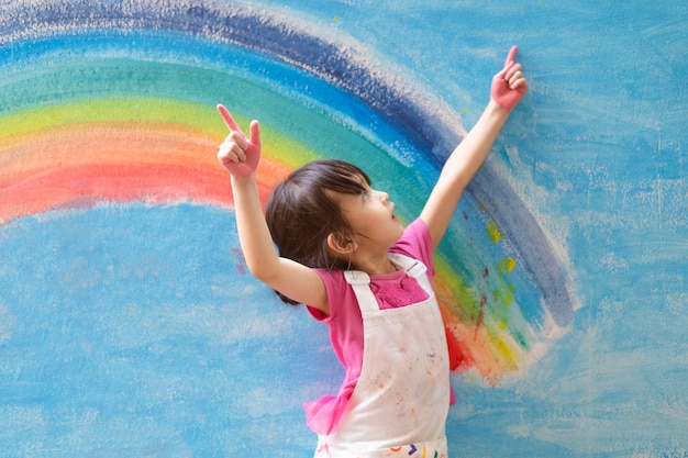 Foto menina asiática está pintando o arco-íris colorido e céu na parede e ela parece feliz e engraçada.