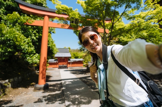 menina asiática feliz de óculos está sorrindo para a câmera enquanto toma selfie com portão torri laranja e santuário distante em um dia ensolarado no templo Tenryuji no japão