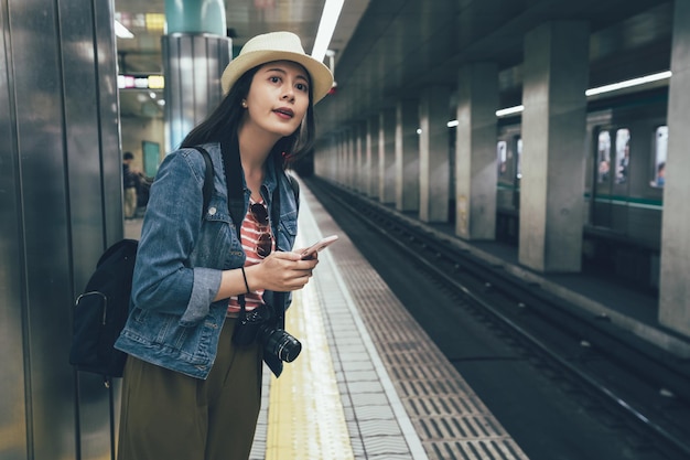 Menina asiática feliz em mensagens de texto de chapéu de palha no telefone inteligente na estação de trem enquanto espera na plataforma. viajante elegante jovem segurando o celular em pé pelo metrô interior comutar subterrâneo.