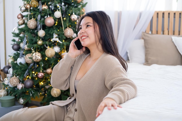 menina asiática feliz em roupas aconchegantes ligando para amigos via celular sentado na cama em casa