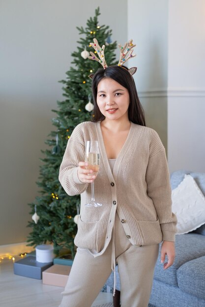 menina asiática feliz em roupas aconchegantes, segurando a taça de champanhe em casa, comemorando o ano novo