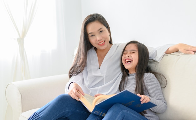 Menina asiática feliz lendo um livro na sala de estar com a mãe dela