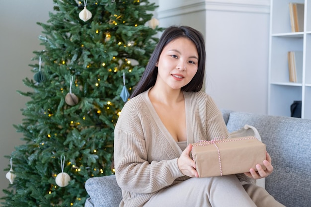 menina asiática feliz no casaco de lã bege com caixa de presente, sentado no sofá em casa perto da árvore de natal