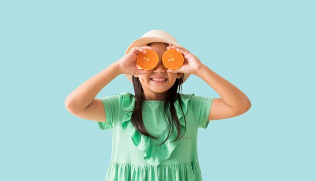 Foto menina asiática feliz posando com um chapéu com óculos de sol segurando fatias de laranja no rosto festa de verão moda vestido verde isolado em fundo azul pastel