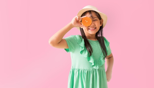 Menina asiática feliz posando com um chapéu com óculos de sol segurando fatias de laranja no rosto Festa de verão moda vestido verde isolado em fundo rosa pastel