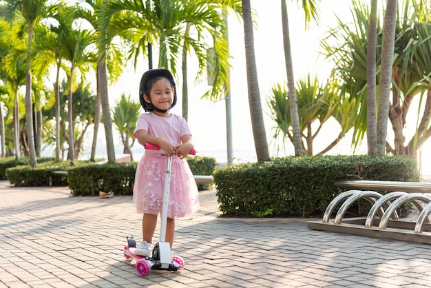 Menina asiática feliz usa capacete seguro jogando prancha rosa na estrada no parque ao ar livre