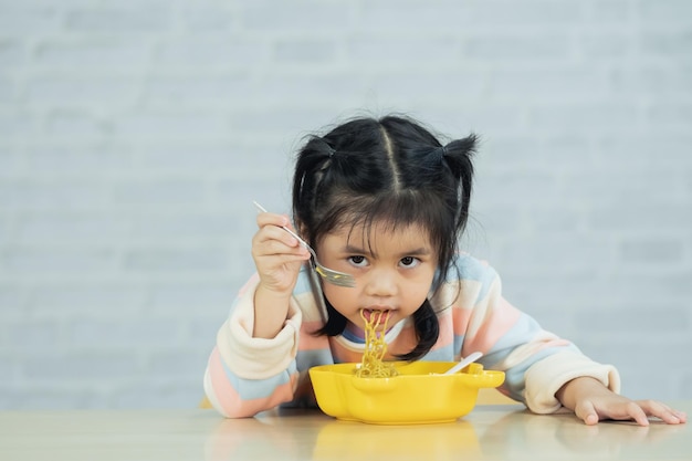 Menina asiática feliz usando colher de talheres e garfo comendo macarrão delicioso e almôndega na cozinha na mesa de jantar Menina asiática feliz praticando comer sozinha na mesa de jantar Conceito de comida para bebê