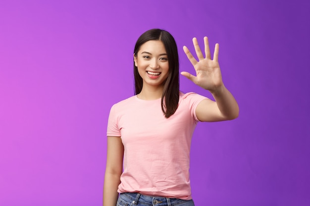 Menina asiática fofa simpática levantando a palma da mão acenando adeus, sorrindo alegremente, adeus, amigos bem-vindos, convidando para entrar, dizendo olá, saudação, fundo roxo