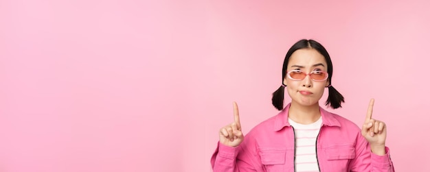 Menina asiática hesitante usa óculos de sol franze a testa e aponta os dedos para cima olha com descrença expressão de rosto cético fica sobre fundo rosa