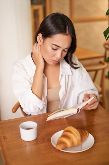 Foto menina asiática lendo livro parecendo triste sentada no café e bebendo café tem um rosto pensativo emburrado