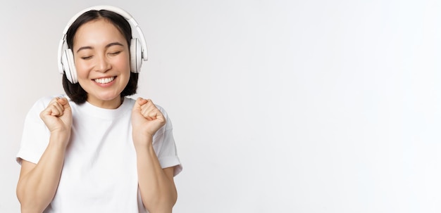 Menina asiática moderna dançando música com fones de ouvido sorrindo feliz em pé na camiseta sobre whit