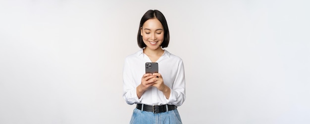 Menina asiática moderna elegante usando aplicativo de celular conversando no celular e sorrindo em pé na blusa branca contra o fundo do estúdio