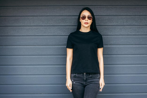 Menina asiática morena elegante vestindo camiseta preta e óculos de sol posando contra o pano urbano de rua