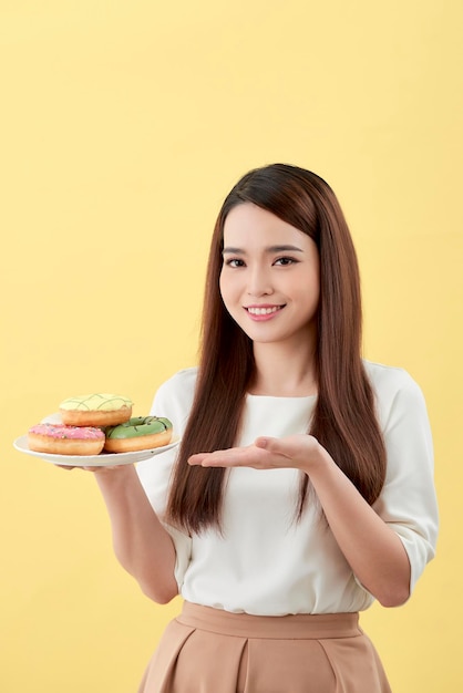 Menina asiática mostra e segurando o prato cheio de saborosos donuts aromáticos deliciosos no rosto de sorriso isolado.