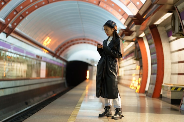 Menina asiática na moda esperando o trem na plataforma do metrô verificar e-mail ou programação online no smartphone