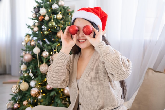 menina asiática no chapéu de papai noel se divertindo, segurando bolas vermelhas de natal, sentado na cama em casa, natal ti