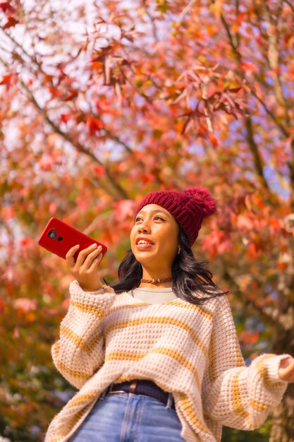 Menina asiática no outono com um celular sorrindo enviando uma mensagem de voz em uma floresta de tecnologia de folhas vermelhas