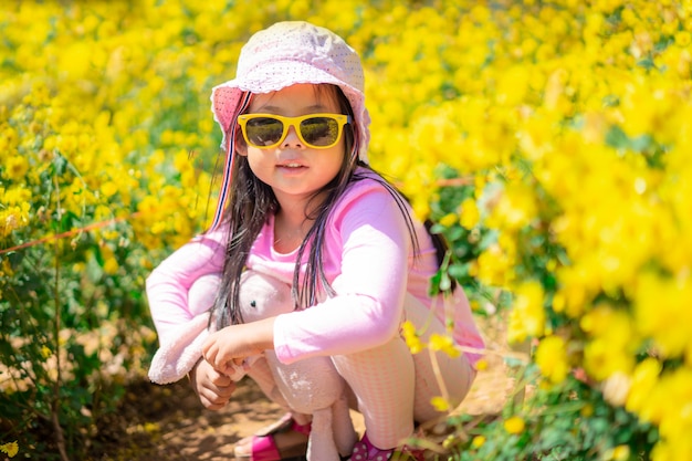 Menina asiática no vestido rosa usar chapéu e óculos de sol, sentado no jardim amarelo