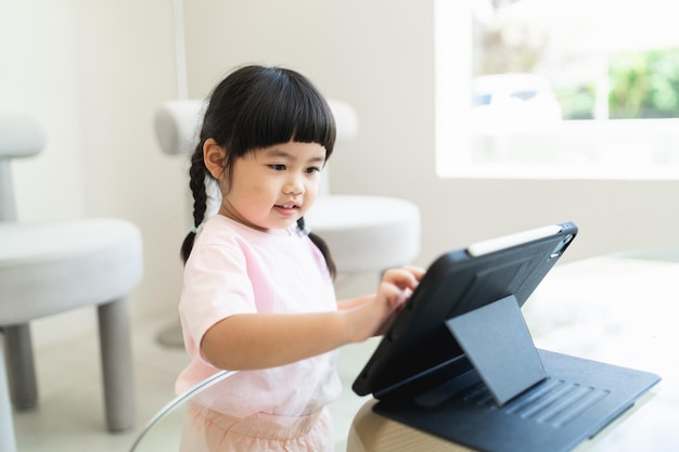 Menina asiática olhando e tocando na tela do tablet com atenção o conceito de crianças superestimuladas Muito tempo na tela menina asiática assistindo vídeos enquanto a tv está funcionando