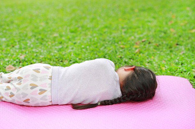 Menina asiática pequena da criança da vista traseira que dorme no colchão cor-de-rosa no gramado da grama verde no jardim do parque do verão.