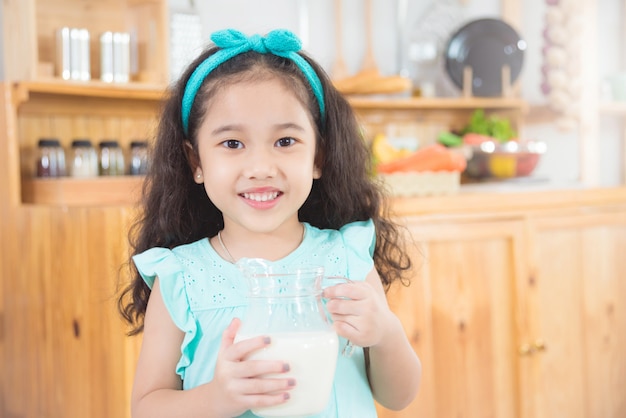 Menina asiática pequena que guarda um jarro de leite e sorri na cozinha