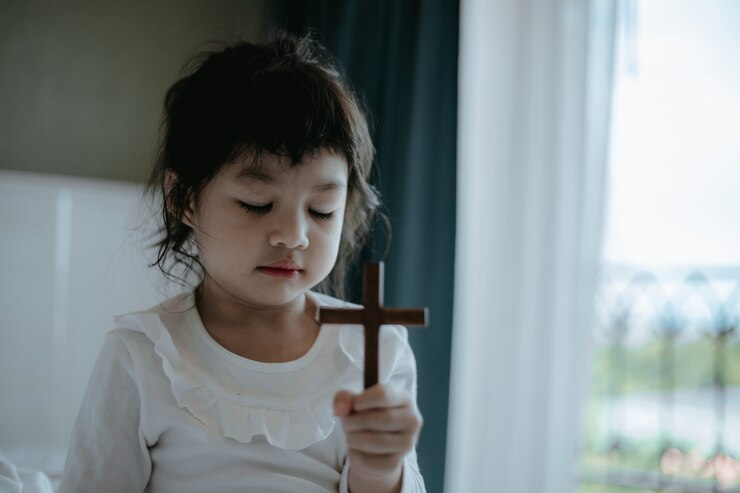 Foto menina asiática pequena que reza e que guarda a cruz de madeira.