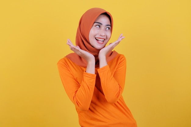 Menina asiática positiva mostrando algo, demonstra espaço de cópia na parede amarela em branco, tem uma expressão amigável e feliz, vestida casualmente com hijab, posa dentro de casa