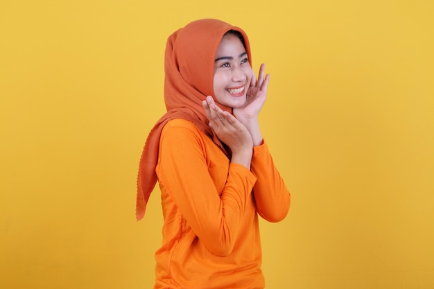 Menina asiática positiva mostrando algo, demonstra espaço de cópia na parede amarela em branco, tem uma expressão amigável e feliz, vestida casualmente com hijab, posa dentro de casa