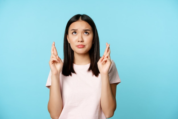 Menina asiática preocupada olhando esperançosa e nervosa, cruza os dedos para dar boa sorte, fazendo um pedido, rezando, usando uma camiseta sobre o fundo azul