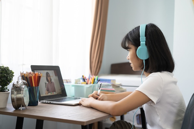 Menina asiática que estuda a lição em linha dos trabalhos de casa em casa, conceito em linha da ideia da educação da distância social