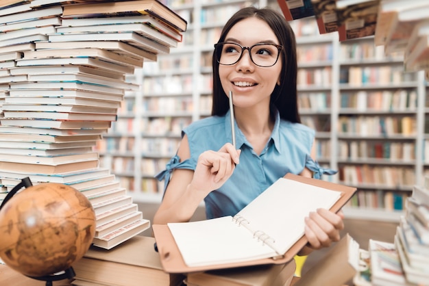 Menina asiática que senta-se na tabela cercada por livros na biblioteca.