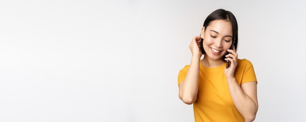 Menina asiática romântica e fofa atende telefonema falando no celular sorrindo coquete em pé em camiseta amarela contra fundo branco