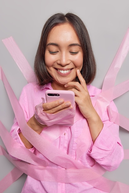 Menina asiática satisfeita da geração do milênio usa informações de pesquisas de celular on-line faz pedidos em pesquisas de celular empregos férias sorrisos feliz usa camisa casual colada na parede cinza