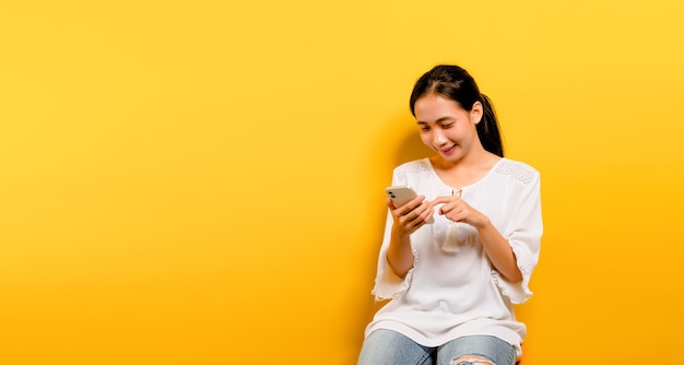 Menina asiática se sentindo feliz, sentada e digitando em um smartphone em um fundo amarelo. Linda asiática