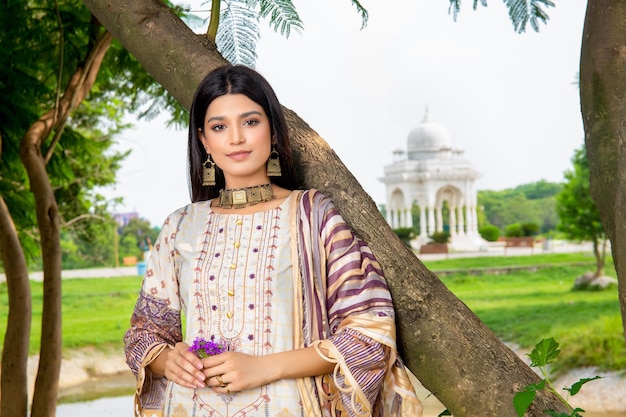 Menina asiática segurando flores e usando joias de vestido Desi tradicionais no jardim com árvore