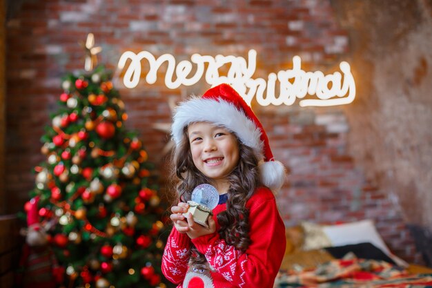 Menina asiática segurando um globo de neve de vidro no fundo de uma árvore de Natal.