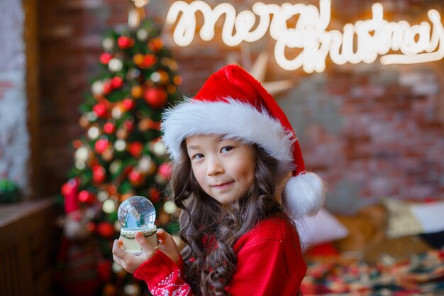 Menina asiática segurando um globo de neve de vidro no fundo de uma árvore de Natal.