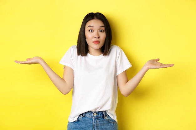 Menina asiática sem noção, encolhendo os ombros, espalhando as mãos e parecendo confusa, não sabe de nada, de pé sobre um fundo amarelo.