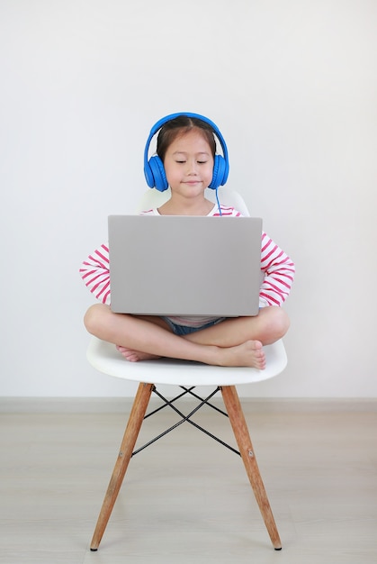 Menina asiática sentada na cadeira usando fone de ouvido, estudo online, aula de aprendizagem por laptop