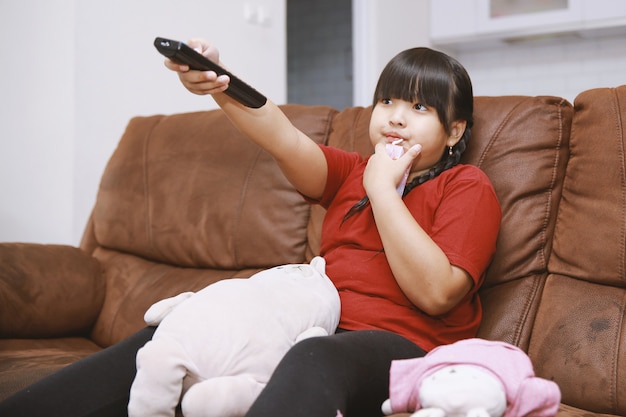 Menina asiática sentada no sofá com o controle remoto assistindo a um filme na televisão na sala de estar