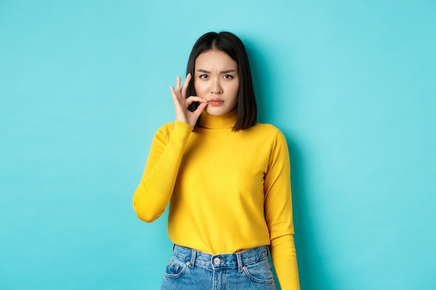 Menina asiática séria mostrando gesto de zíper de boca, promessa de manter a boca fechada e carrancuda, contando o grande segredo, de pé na camisola amarela sobre fundo azul.