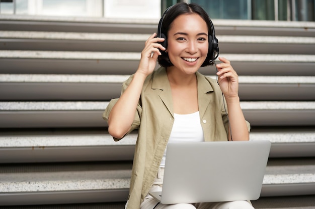 Menina asiática sorridente em fones de ouvido trabalha em laptop nômade digital usando computador em controle remoto sentado em s